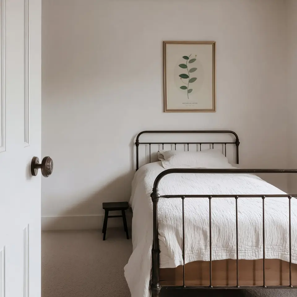Quiet Iron Bed: A small pale gold frame hangs on a white wall above a black iron bed dressed in white linen, seen through an open panelled door with a dark stool at the bedside.